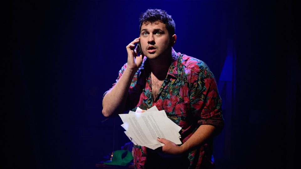 Will Jackson, Yours Sincerely a man with short, dark hair, a Hawaiian shirt and gold painted nails looks up expectantly as he clutches some papers and brings his mobile phone to his ear