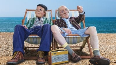 Dead Good - Characters Bob and Bernard (people wearing realistic style masks) Bob & Bernard sit in their deck chairs on a pebble beach looking out, the sea is behind them and they have a radio between them.