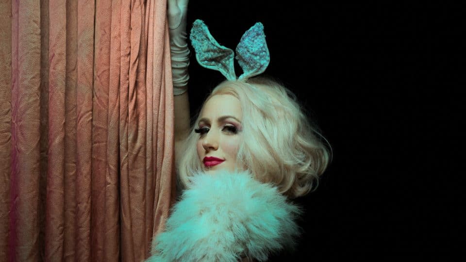 Hundred Watt Club - an image of Lena wearing bunny ears and a feather boa, looking over her shoulder towards camera, her gloved hand against a wall