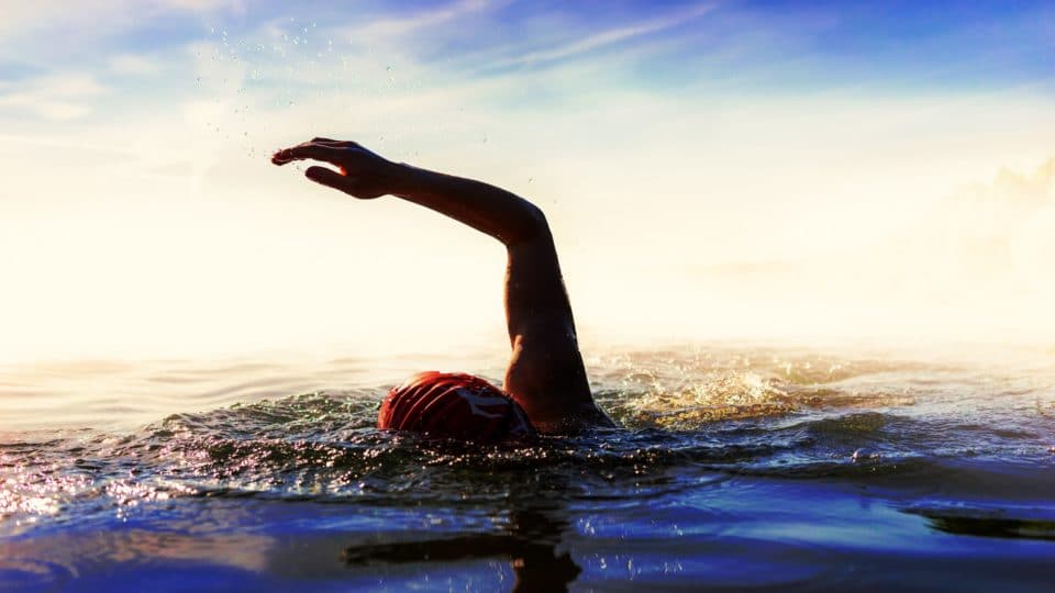 When the Long Trick's Over - image of someone swimming in the sea with an arm raised over their head as they take a stroke. They are wearing a red swimming cap and the sky is in the background