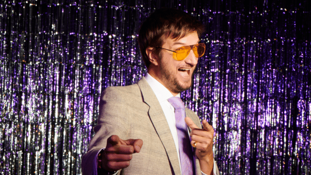 Showman in sunglasses pointing at screen in front of glitter curtain