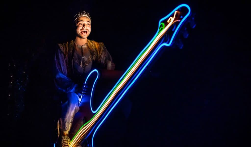Panto Heroes - Picture of James Hameed as Aladdin riding his guitar which is lit up with neon lights