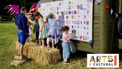 A family create colourful artwork on the side of a caravan on a field surrounded by haybales and colourful standards. A vibrant logo for the Arts and Cultural fund is in the bottom right-hand corner.