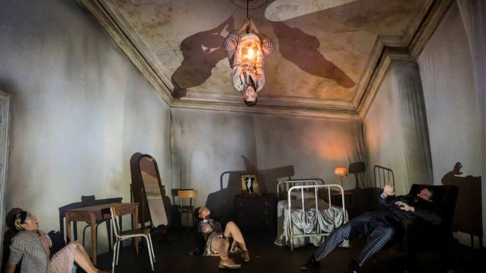 man hanging upside down on the hanging light while two women and another man on the floor looking at him scared