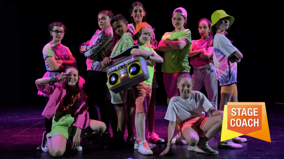 A crowd of young people dressed in neon pinks, greens, and oranges pose with an inflatable boombox and a whole lot of attitude with a Stagecoach Logo in the bottom right-hand corner.