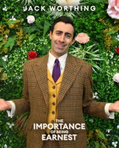 Jack Worthing in suit shrugging comedically in front of a floral wall
