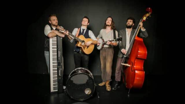 Four men each holding a keyboard, a guitar, a banjo and a cello respectively with a drum placed in the front