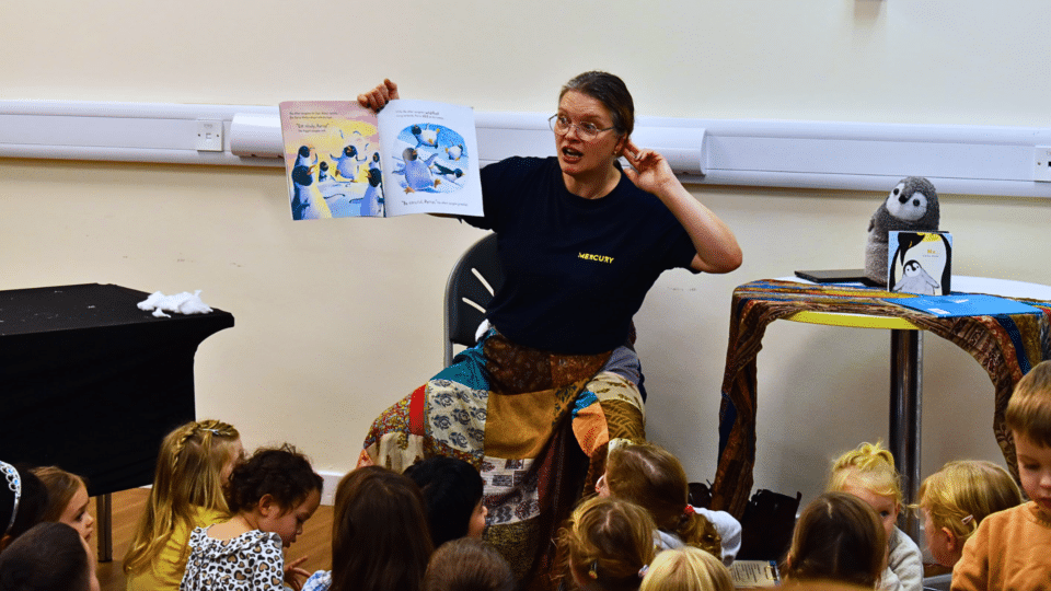 Kate Milner reading a children's story book to a group of young children sat on the floor in the library