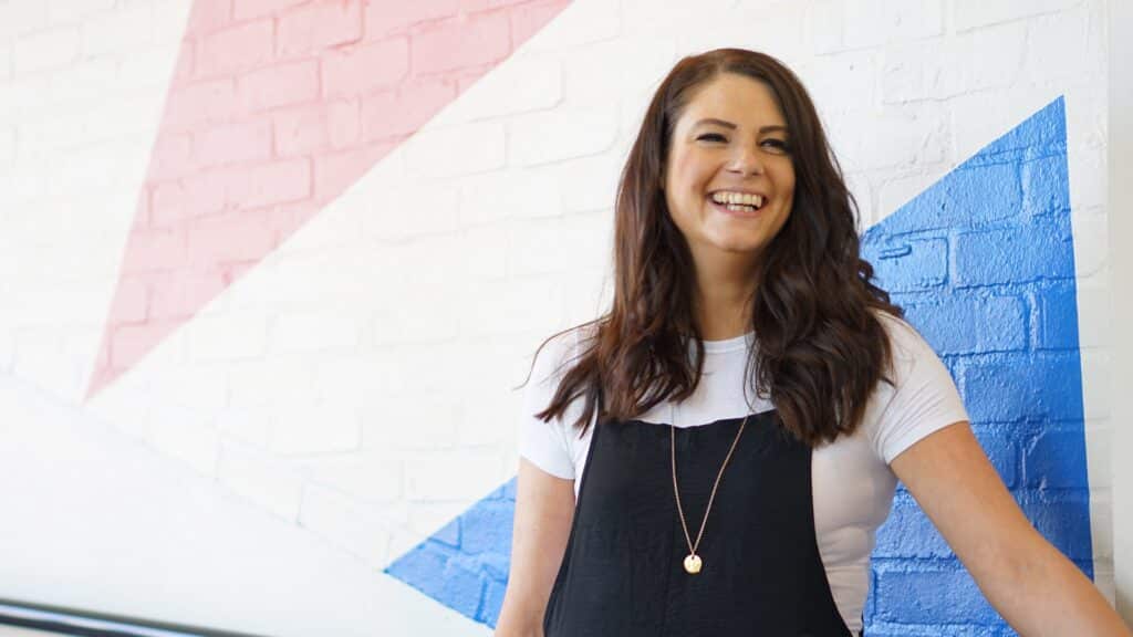 Natasha Rickman 1 1920x1080 Photo of Natasha Rickman, a white woman with brown hair, smiling looking past the camera, against a white wall with pink and blue triangles.