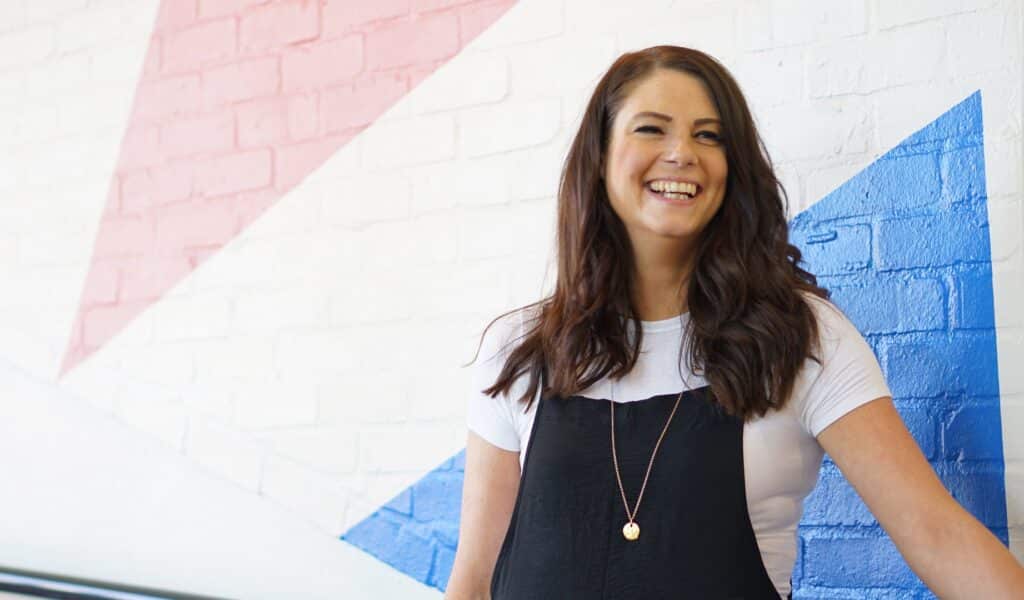 Photo of Natasha Rickman, a white woman with brown hair, smiling looking past the camera, against a white wall with pink and blue triangles.